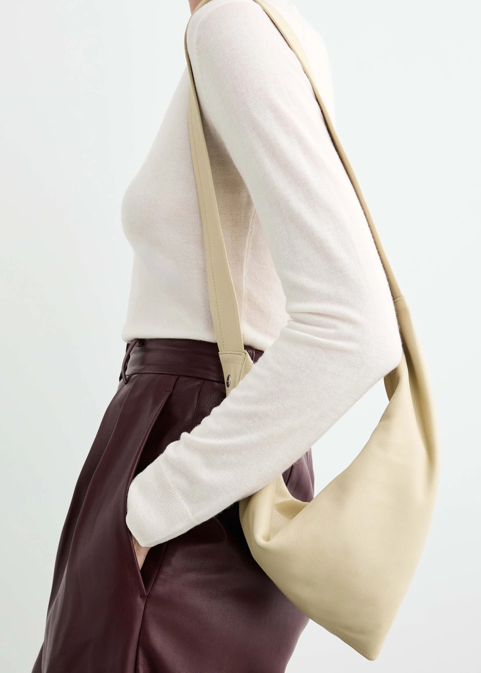 A person in a white long-sleeve top and high-waisted burgundy pants carries a soft, beige Vancouver Sling Bag. The image is cropped to show only the upper body, focusing on the outfit and accessories.