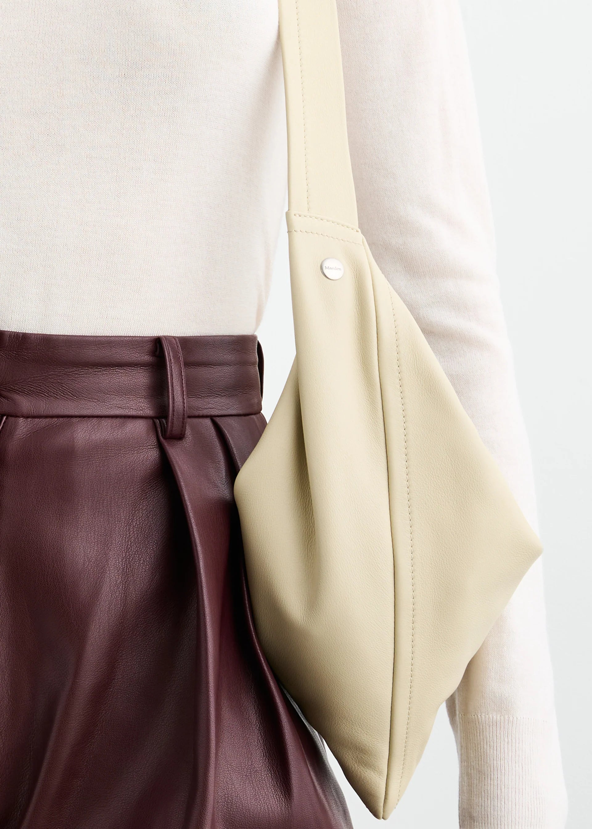 A person wearing a cream long-sleeve top, high-waisted maroon leather pants, and carrying the Vancouver Sling Bag—a sculptural shoulder bag crafted from nappa leather.