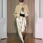 A model walks on a wooden floor in front of beige curtains and white double doors, wearing Zoela Pants—flowing cream and gray pants with a barrel-leg silhouette—styled with a beige high-collar jacket and a white top adorned with gold embellishment.