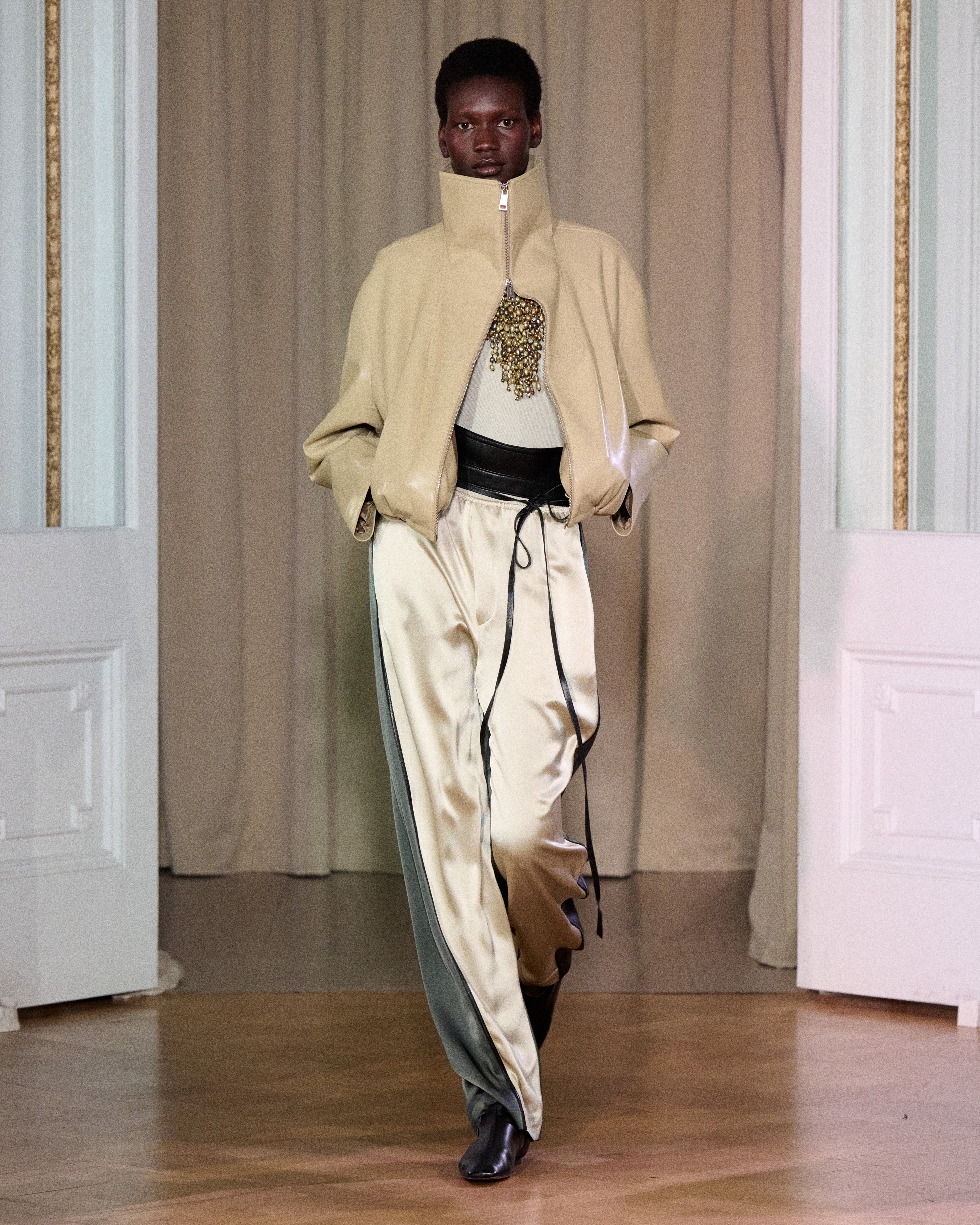 A model walks on a wooden floor in front of beige curtains and white double doors, wearing Zoela Pants—flowing cream and gray pants with a barrel-leg silhouette—styled with a beige high-collar jacket and a white top adorned with gold embellishment.