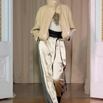 A model walks on a wooden floor in front of beige curtains and white double doors, wearing Zoela Pants—flowing cream and gray pants with a barrel-leg silhouette—styled with a beige high-collar jacket and a white top adorned with gold embellishment.