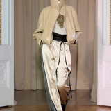 A model walks on a wooden floor in front of beige curtains and white double doors, wearing Zoela Pants—flowing cream and gray pants with a barrel-leg silhouette—styled with a beige high-collar jacket and a white top adorned with gold embellishment.