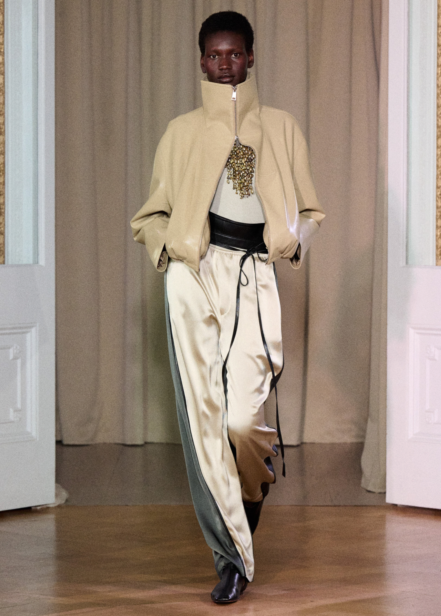 A model walks on a wooden floor in front of beige curtains and white double doors, wearing Zoela Pants—flowing cream and gray pants with a barrel-leg silhouette—styled with a beige high-collar jacket and a white top adorned with gold embellishment.