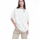 Wearing the Relaxed Fit T-Shirt with Lace Overlayer, a person with long, straight dark hair stands against a white background with one hand in their pocket, styled in beige wide-leg pants.