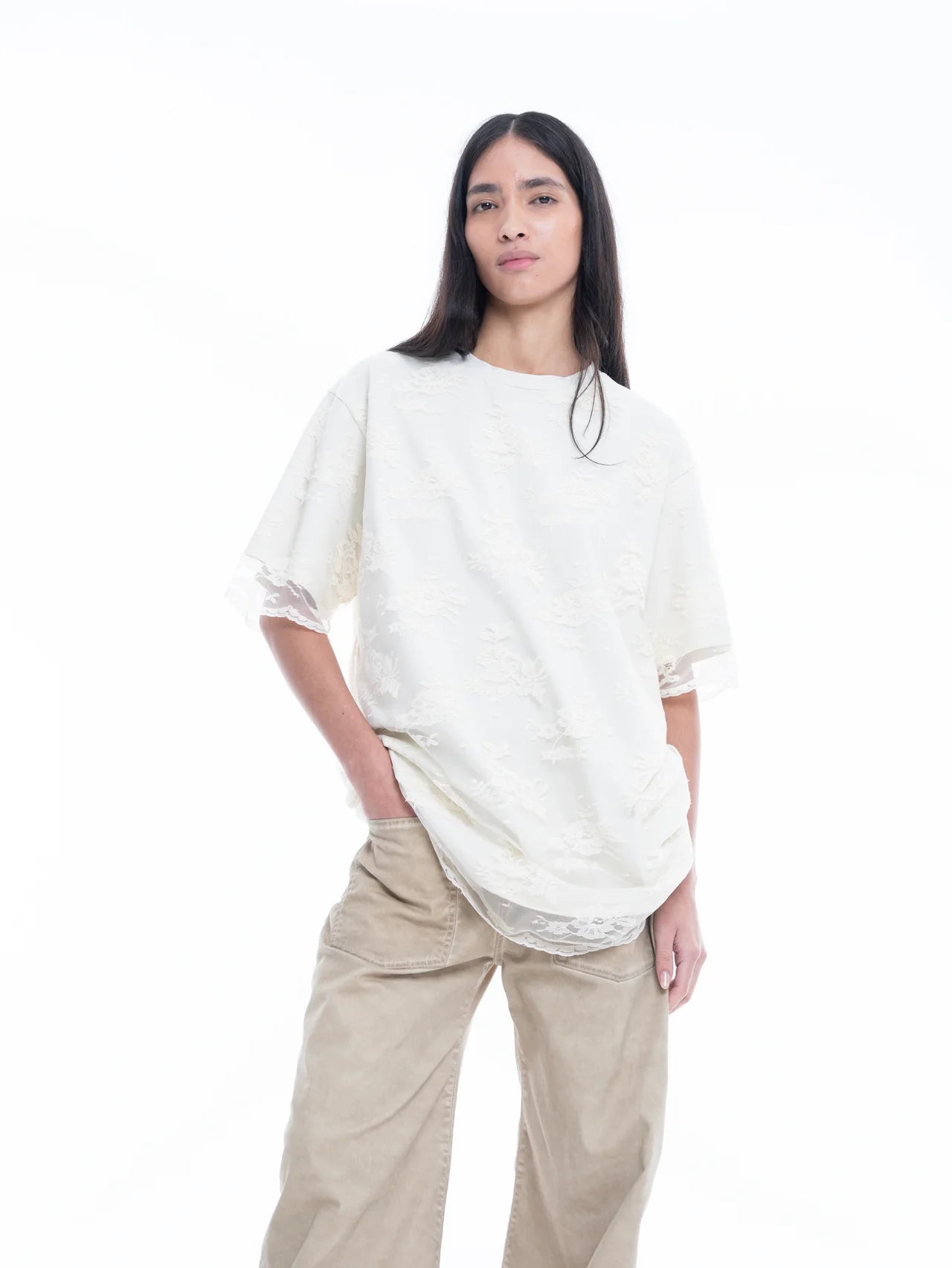 Wearing the Relaxed Fit T-Shirt with Lace Overlayer, a person with long, straight dark hair stands against a white background with one hand in their pocket, styled in beige wide-leg pants.