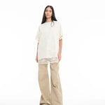 A person with long dark hair wears the Relaxed Fit T-Shirt with Lace Overlayer in off-white, styled over beige wide-leg pants and green sandals, standing against a plain white background.