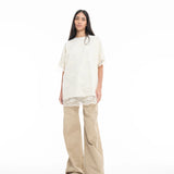 A person with long dark hair wears the Relaxed Fit T-Shirt with Lace Overlayer in off-white, styled over beige wide-leg pants and green sandals, standing against a plain white background.