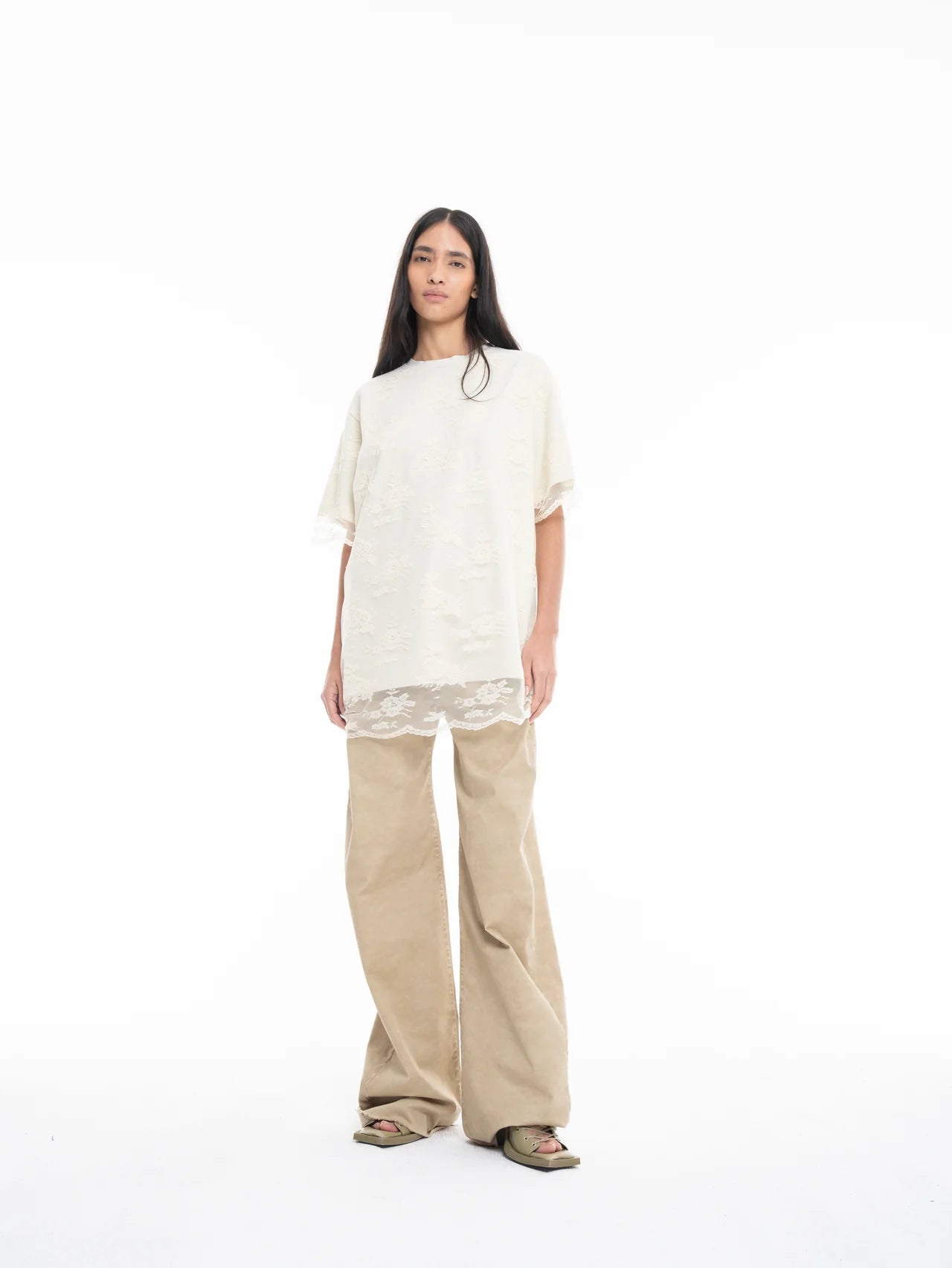 A person with long dark hair wears the Relaxed Fit T-Shirt with Lace Overlayer in off-white, styled over beige wide-leg pants and green sandals, standing against a plain white background.