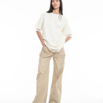 A woman stands against a white background, wearing the Relaxed Fit T-Shirt with Lace Overlayer in off-white, wide-legged beige pants, and open-toe sandals. She has long dark hair and her hands in her pockets.