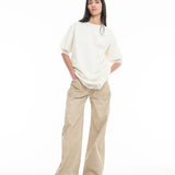 A woman stands against a white background, wearing the Relaxed Fit T-Shirt with Lace Overlayer in off-white, wide-legged beige pants, and open-toe sandals. She has long dark hair and her hands in her pockets.
