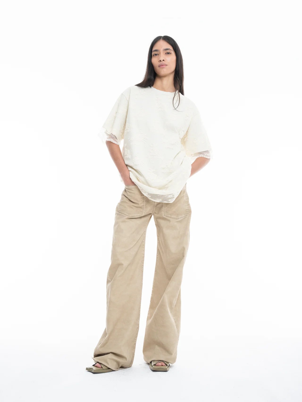 A woman stands against a white background, wearing the Relaxed Fit T-Shirt with Lace Overlayer in off-white, wide-legged beige pants, and open-toe sandals. She has long dark hair and her hands in her pockets.