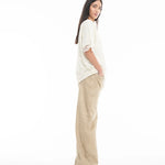 A woman with long brown hair stands sideways, wearing a Relaxed Fit T-Shirt with Lace Overlayer and wide-leg tan pants, hands in pockets, set against a plain white background.
