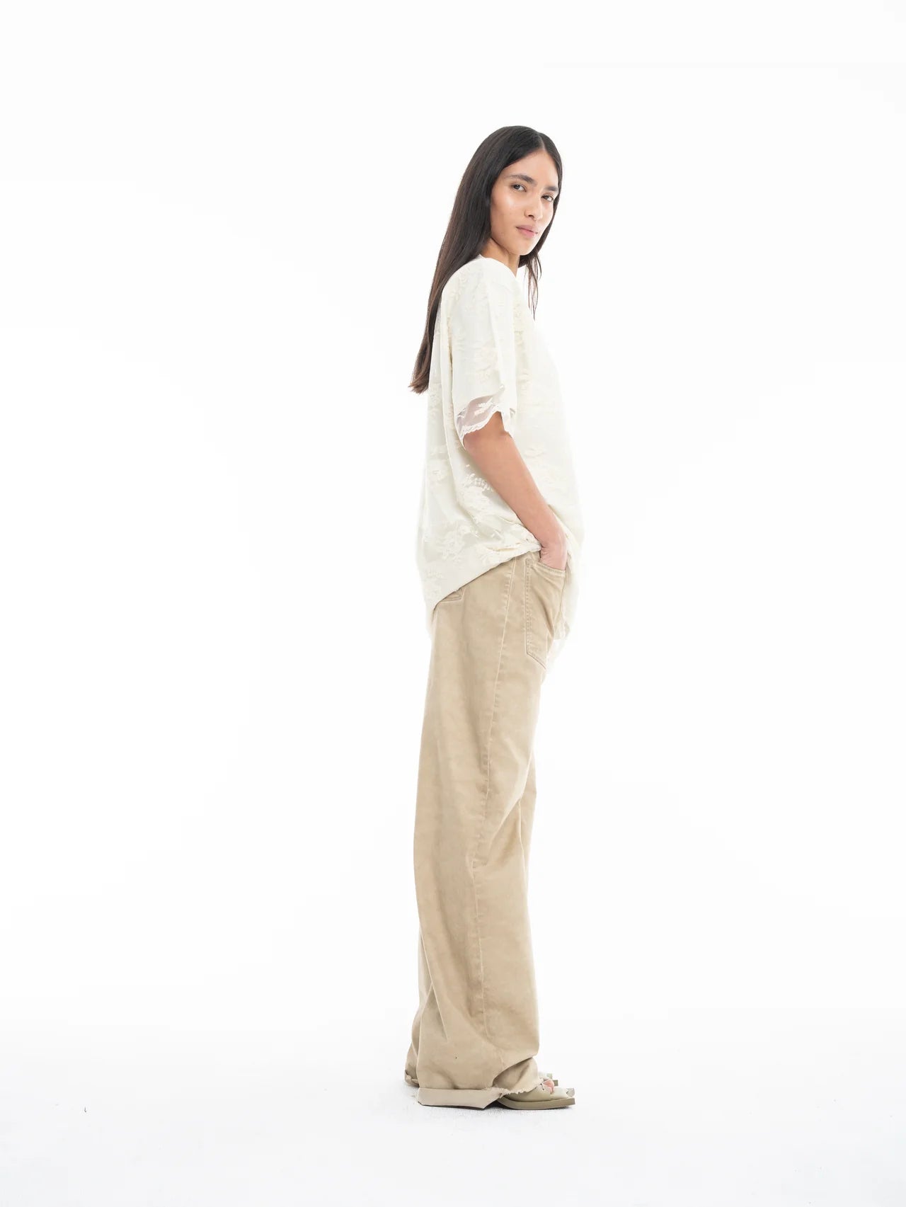 A woman with long brown hair stands sideways, wearing a Relaxed Fit T-Shirt with Lace Overlayer and wide-leg tan pants, hands in pockets, set against a plain white background.