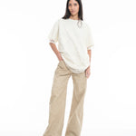 A woman with long dark hair stands against a white background, wearing the Relaxed Fit T-Shirt with Lace Overlayer, paired with wide-legged beige pants and green sandals. She has one hand in her pocket and a neutral expression.