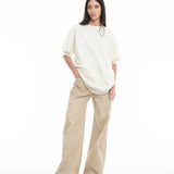 A woman with long dark hair stands against a white background, wearing the Relaxed Fit T-Shirt with Lace Overlayer, paired with wide-legged beige pants and green sandals. She has one hand in her pocket and a neutral expression.