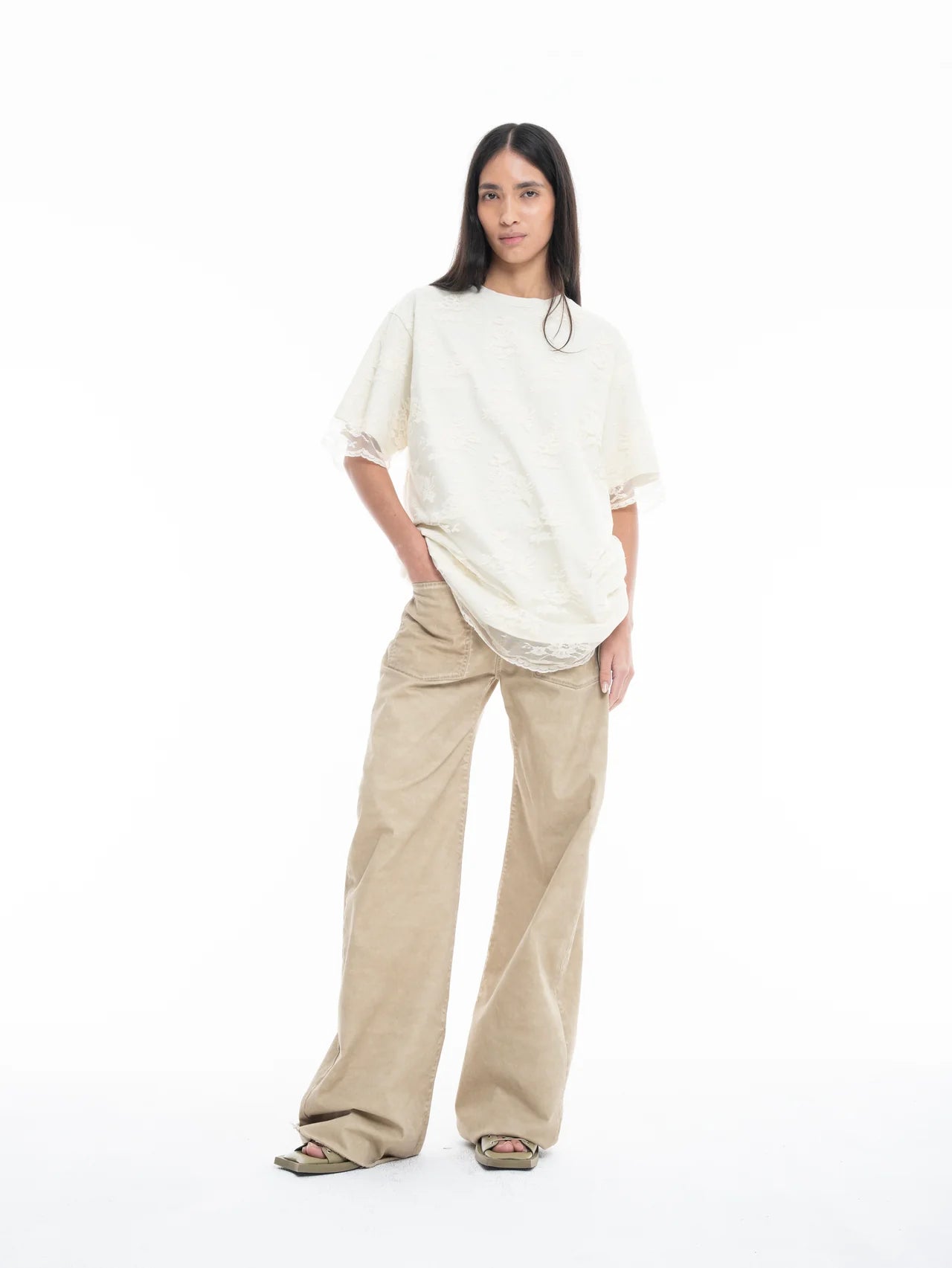 A woman with long dark hair stands against a white background, wearing the Relaxed Fit T-Shirt with Lace Overlayer, paired with wide-legged beige pants and green sandals. She has one hand in her pocket and a neutral expression.