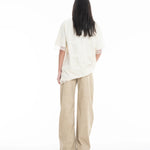 A person with long dark hair, wearing the Relaxed Fit T-Shirt with Lace Overlayer and wide-legged beige pants, stands facing away against a plain white background.