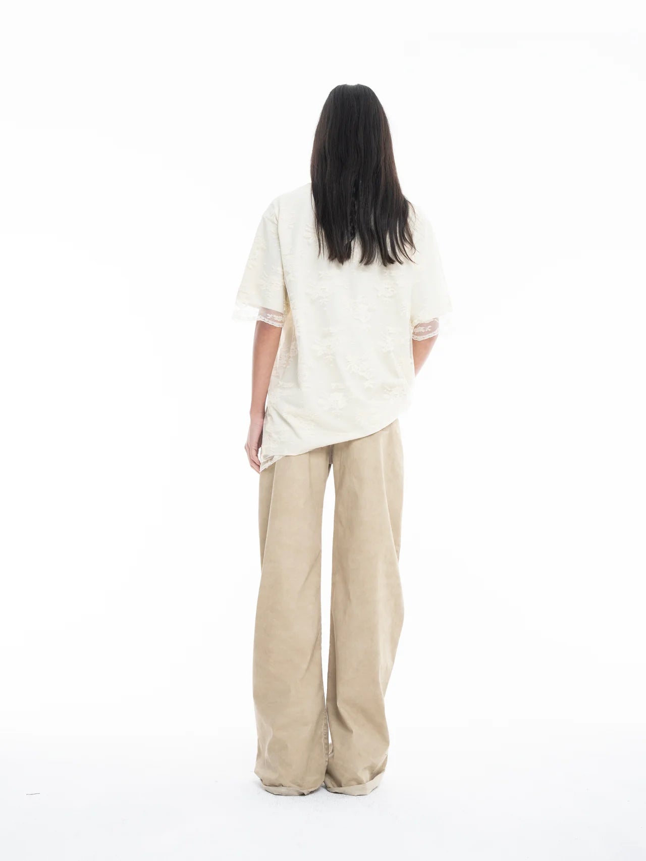A person with long dark hair, wearing the Relaxed Fit T-Shirt with Lace Overlayer and wide-legged beige pants, stands facing away against a plain white background.