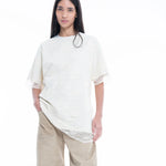 A person with long dark hair wears the Relaxed Fit T-Shirt with Lace Overlayer in an elegant off-white shade, paired with beige wide-leg pants, standing against a plain white background.