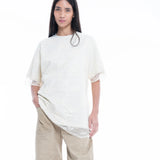 A person with long dark hair wears the Relaxed Fit T-Shirt with Lace Overlayer in an elegant off-white shade, paired with beige wide-leg pants, standing against a plain white background.