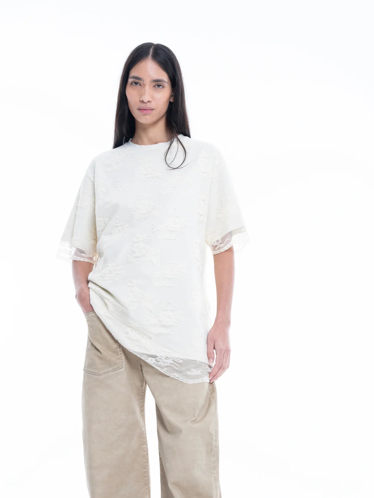 A person with long dark hair wears the Relaxed Fit T-Shirt with Lace Overlayer in an elegant off-white shade, paired with beige wide-leg pants, standing against a plain white background.