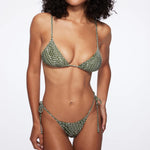A woman wearing the Praia Bottom, a green patterned triangle bikini bottom with adjustable side ties, paired with sunglasses and a woven bucket hat, poses against a plain white background.
