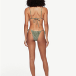 A woman stands barefoot facing away from the camera against a plain white background, wearing the green patterned Praia Bottom bikini with adjustable side ties and a straw hat.