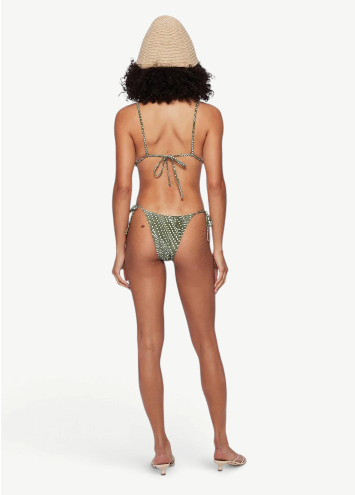 A woman stands barefoot facing away from the camera against a plain white background, wearing the green patterned Praia Bottom bikini with adjustable side ties and a straw hat.