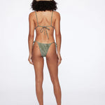 A woman stands barefoot facing away from the camera against a plain white background, wearing the green patterned Praia Bottom bikini with adjustable side ties and a straw hat.