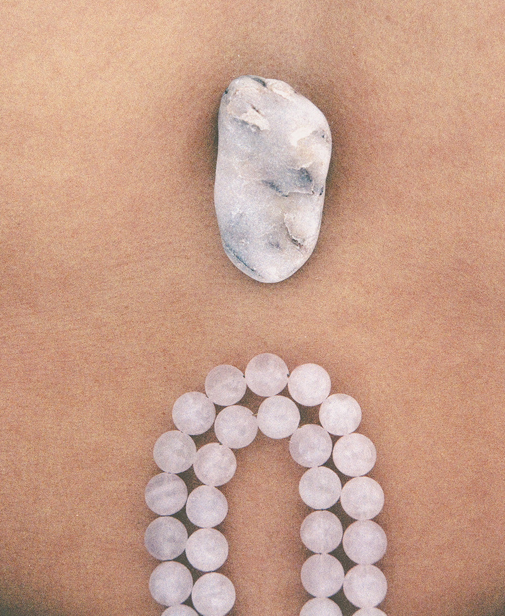 A smooth, oval stone rests on a tan surface above a loop of round, translucent beads arranged in a curve, reminiscent of the delicately handcrafted Petra Straps in pink quartz from Paris.