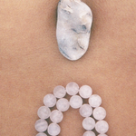 A smooth, oval stone rests on a tan surface above a loop of round, translucent beads arranged in a curve, reminiscent of the delicately handcrafted Petra Straps in pink quartz from Paris.
