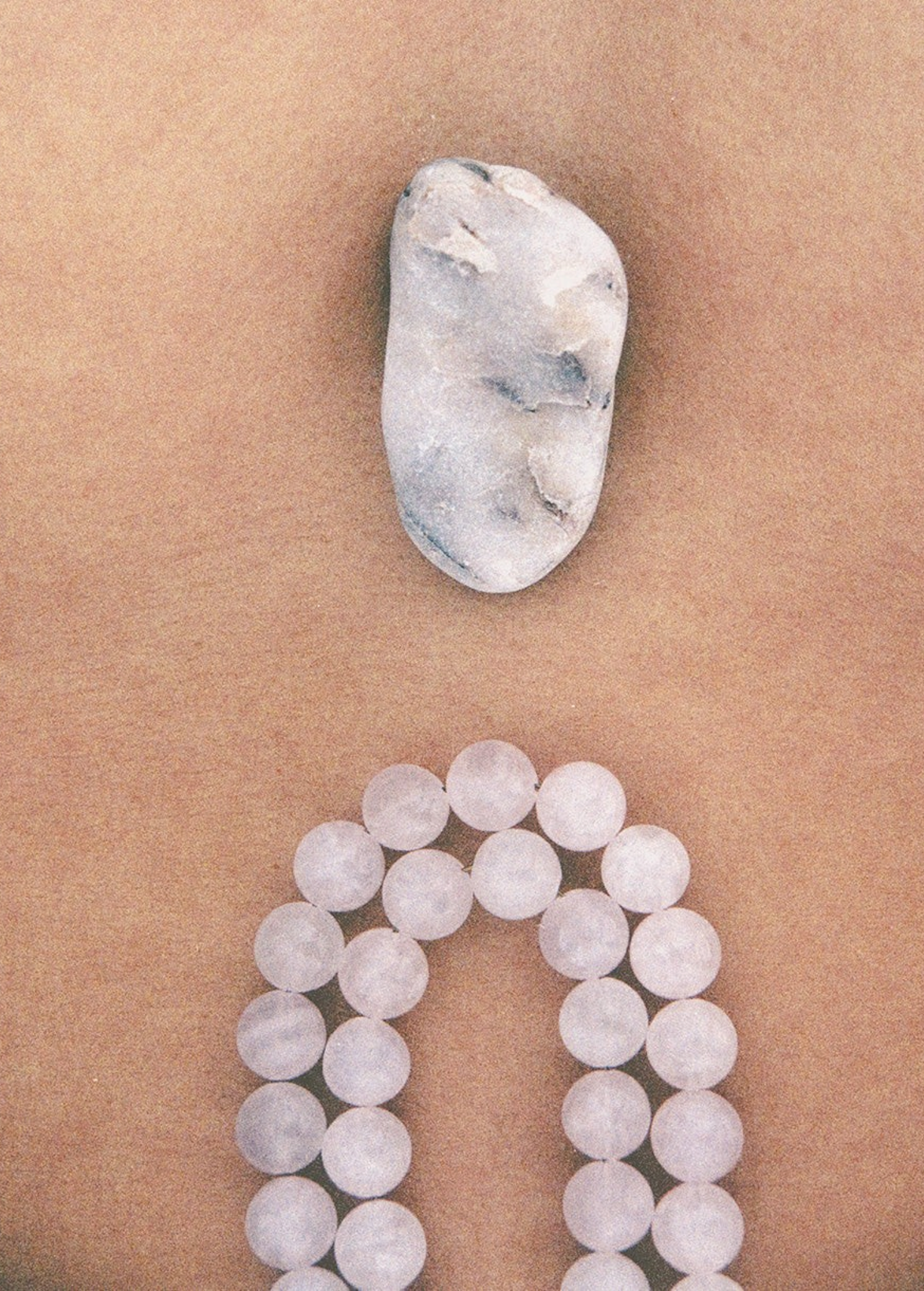 A smooth, oval stone rests on a tan surface above a loop of round, translucent beads arranged in a curve, reminiscent of the delicately handcrafted Petra Straps in pink quartz from Paris.