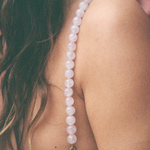 A close-up of a woman’s shoulder displays a Petra Strap, handmade in Paris, featuring delicate pale pink quartz beads draped over her white top. Her wavy brown hair cascades down her back against a softly blurred background.