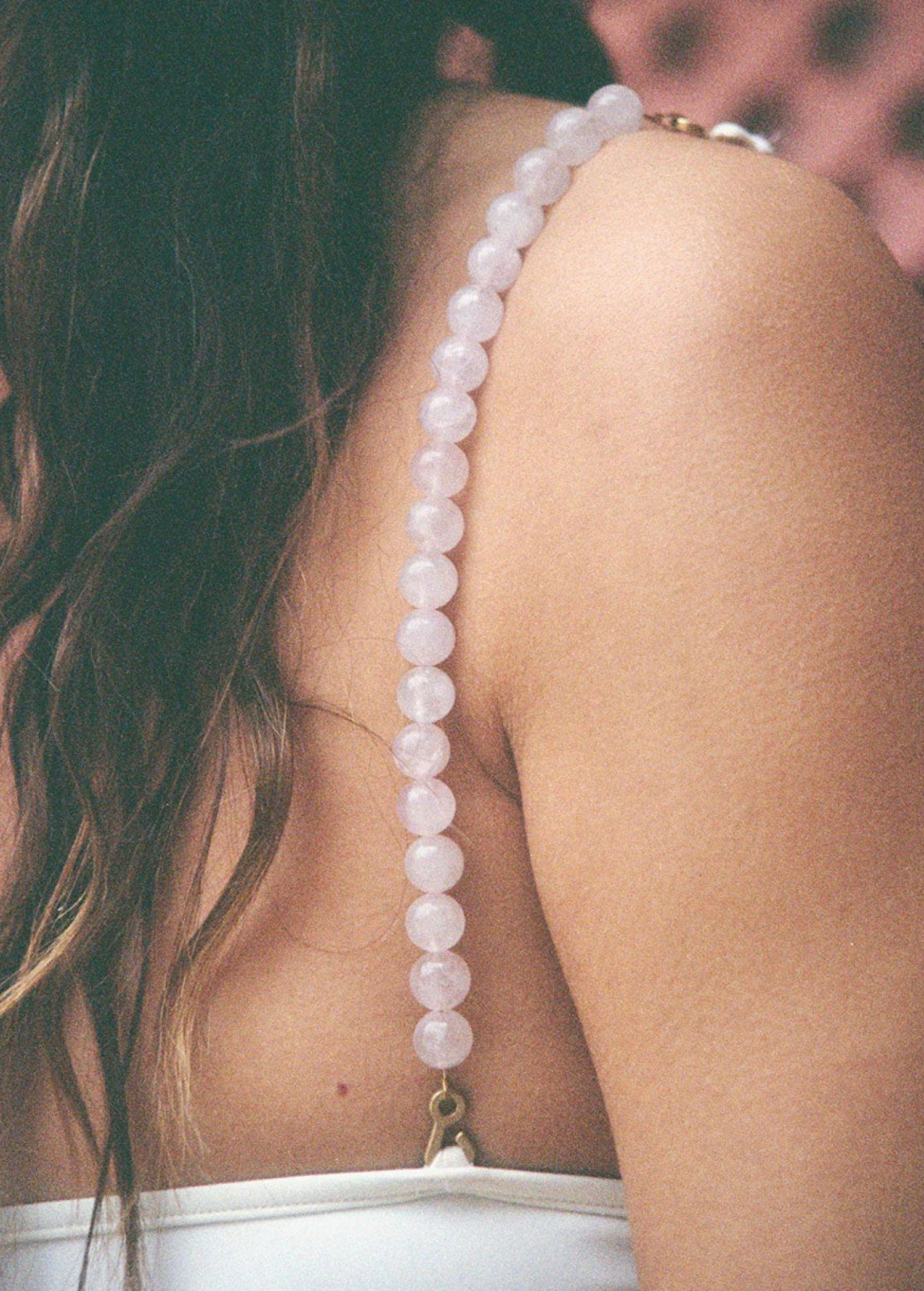 A close-up of a woman’s shoulder displays a Petra Strap, handmade in Paris, featuring delicate pale pink quartz beads draped over her white top. Her wavy brown hair cascades down her back against a softly blurred background.