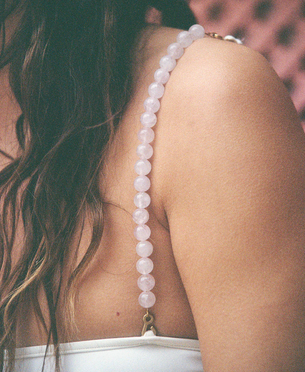 A close-up of a woman’s shoulder displays a Petra Strap, handmade in Paris, featuring delicate pale pink quartz beads draped over her white top. Her wavy brown hair cascades down her back against a softly blurred background.