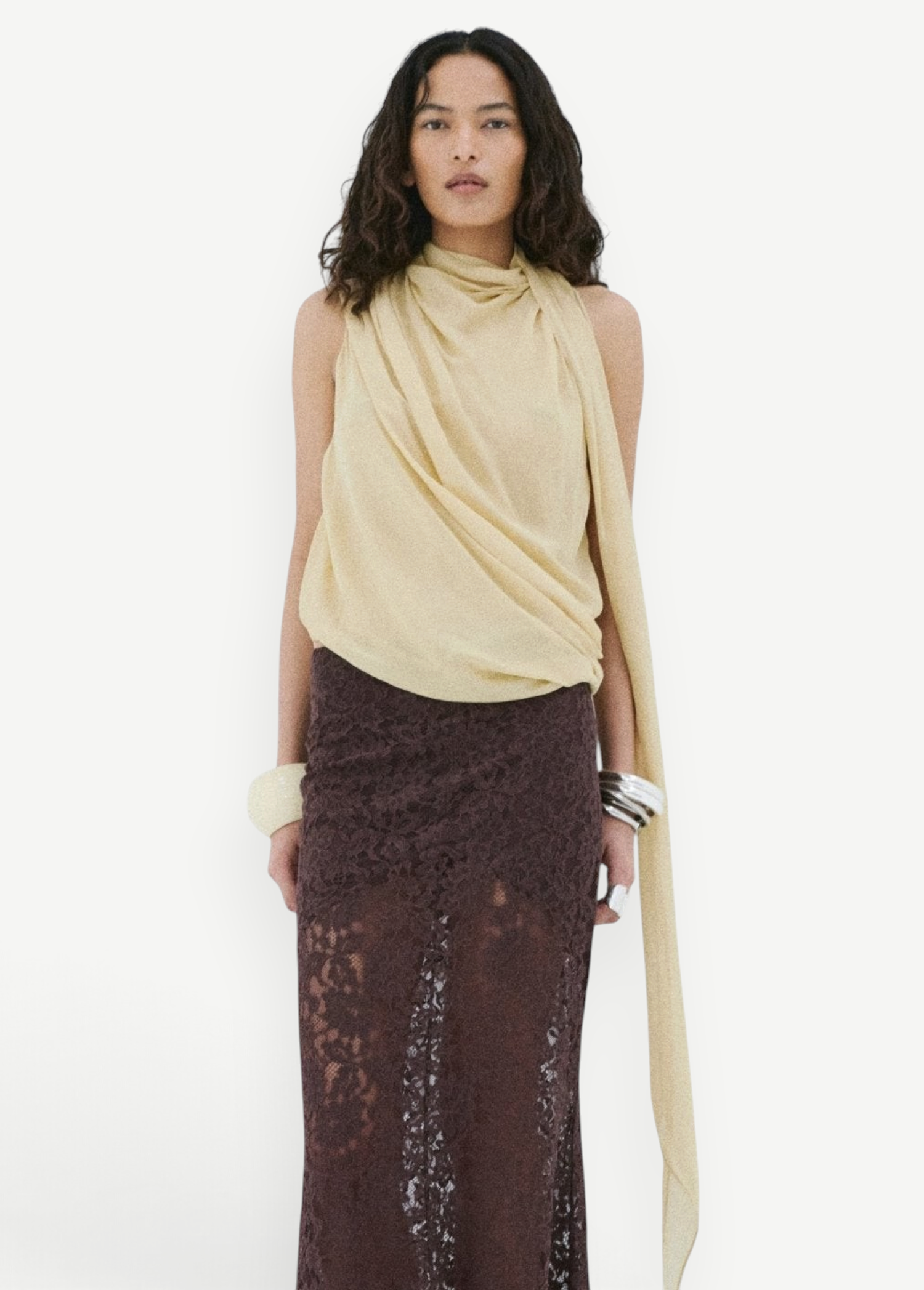A model wearing a pale yellow Menorca top with an asymmetric silhouette and a floor-length brown lace skirt.
