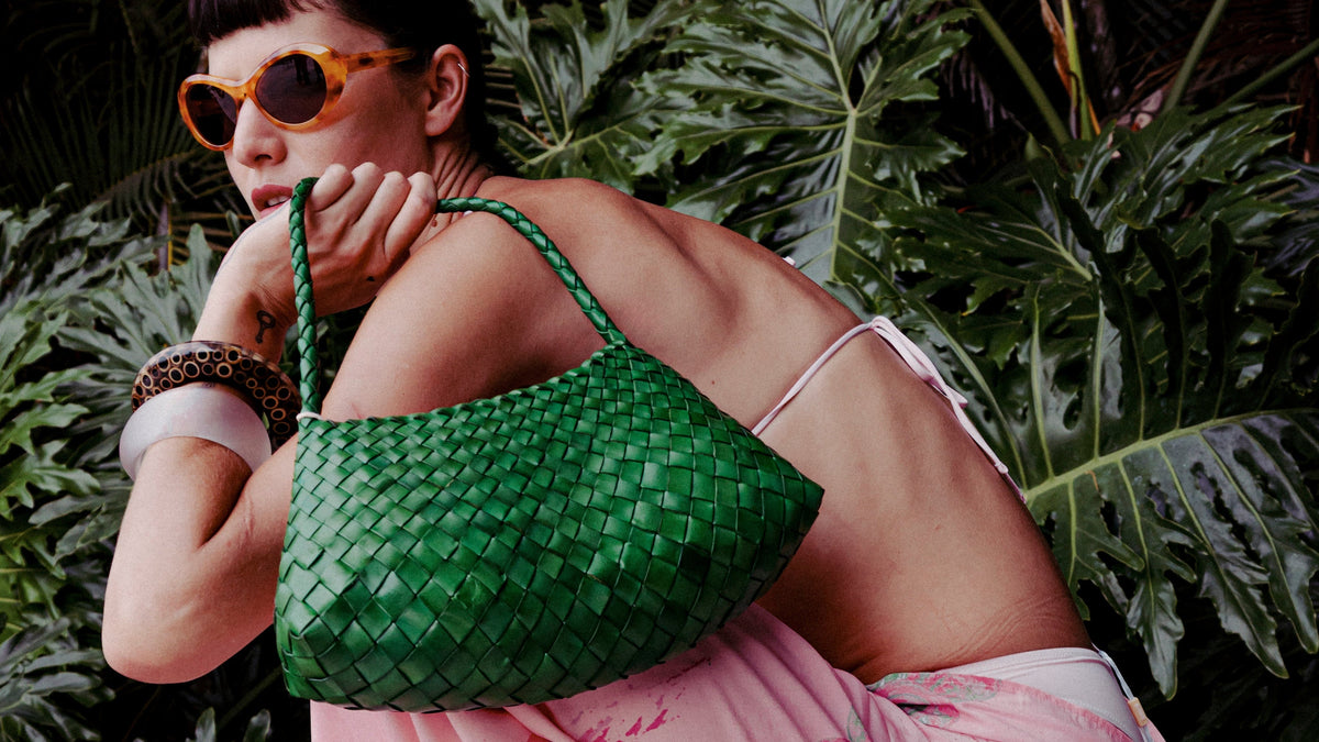 A woman in a pink paisley dress and orange-tinted sunglasses carries a green woven shoulder bag, posed outdoors against a backdrop of lush tropical green foliage.