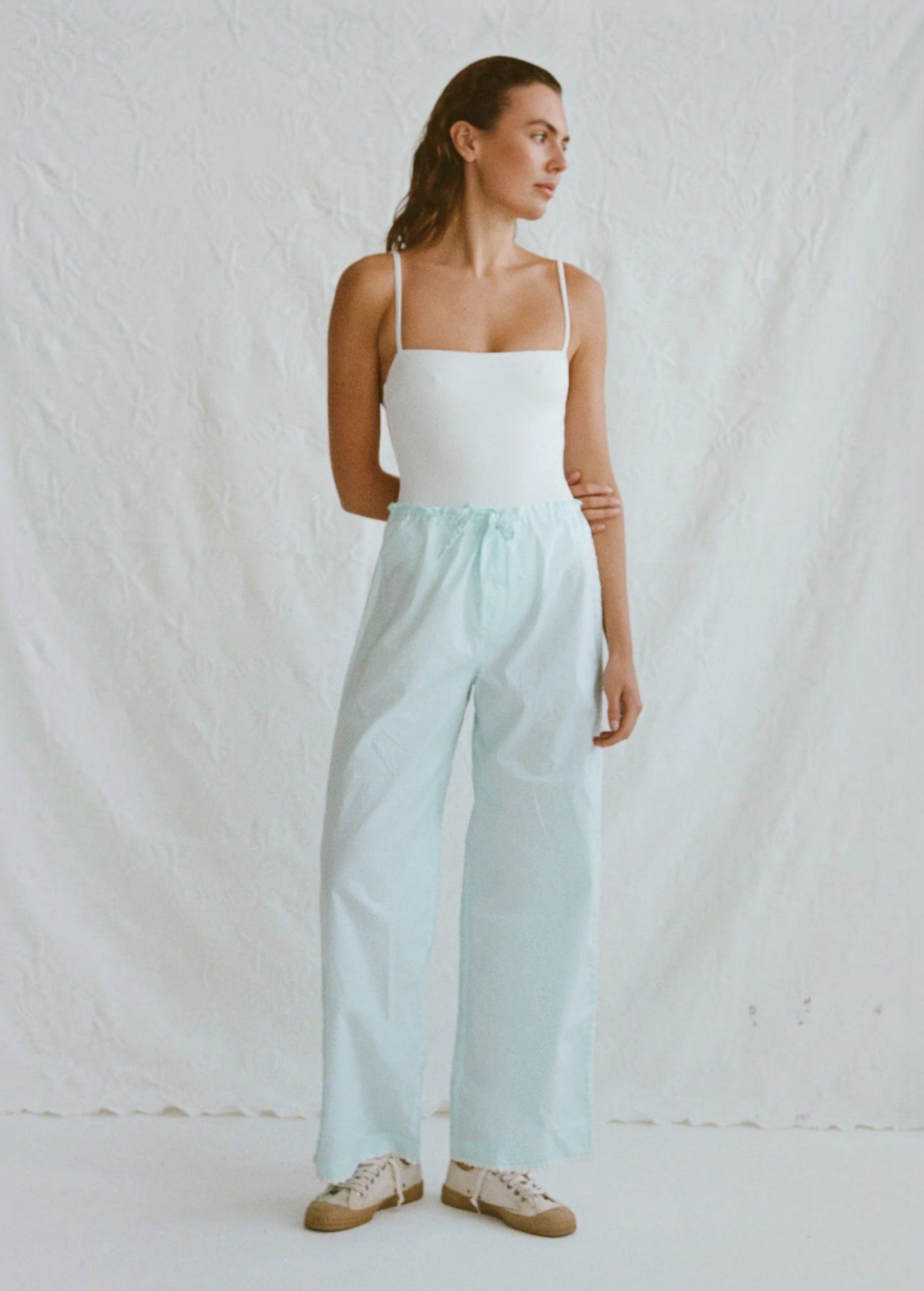 A woman wearing a white camisole and light blue organic cotton Casa Pant wide-leg pants with white sneakers.