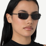 A woman with long black hair wears narrow, rectangular black 2000s sunglasses and a sleeveless black top, standing against a plain white background and effortlessly accessorizing her look with the chic designer Paris handbag.