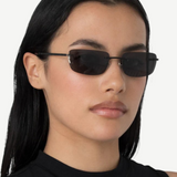 A woman with long black hair wears narrow, rectangular black 2000s sunglasses and a sleeveless black top, standing against a plain white background and effortlessly accessorizing her look with the chic designer Paris handbag.