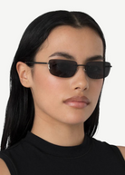 A woman with long black hair wears narrow, rectangular black 2000s sunglasses and a sleeveless black top, standing against a plain white background and effortlessly accessorizing her look with the chic designer Paris handbag.
