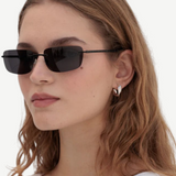 A woman with wavy brown hair wears the Paris black rectangular 2000s sunglasses, silver hoop earrings, and a white tank top, standing against a plain light background.