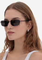 A woman with wavy brown hair wears the Paris black rectangular 2000s sunglasses, silver hoop earrings, and a white tank top, standing against a plain light background.