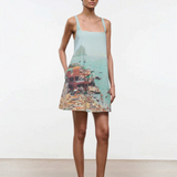 A woman wears a light blue, sleeveless Brio Dress printed with a coastal beach town scene, styled as a versatile day-to-night essential.