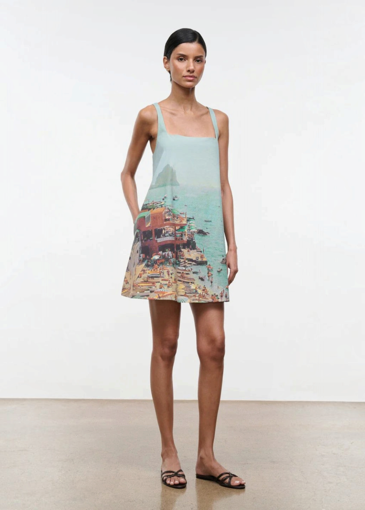 A woman wears a light blue, sleeveless Brio Dress printed with a coastal beach town scene, styled as a versatile day-to-night essential.