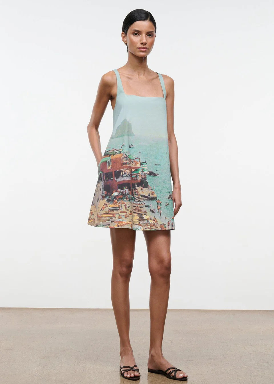 A woman wears a light blue, sleeveless Brio Dress printed with a coastal beach town scene, styled as a versatile day-to-night essential.