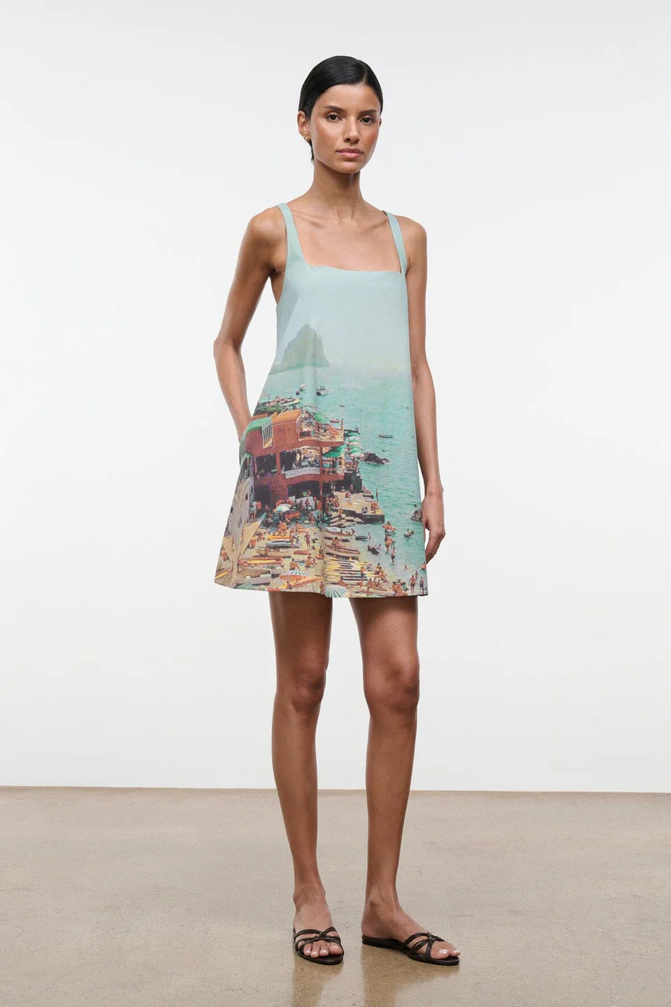 A woman wears a light blue, sleeveless Brio Dress printed with a coastal beach town scene, styled as a versatile day-to-night essential.
