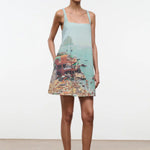 A woman wears a light blue, sleeveless Brio Dress printed with a coastal beach town scene, styled as a versatile day-to-night essential.