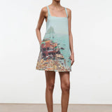 A woman wears a light blue, sleeveless Brio Dress printed with a coastal beach town scene, styled as a versatile day-to-night essential.