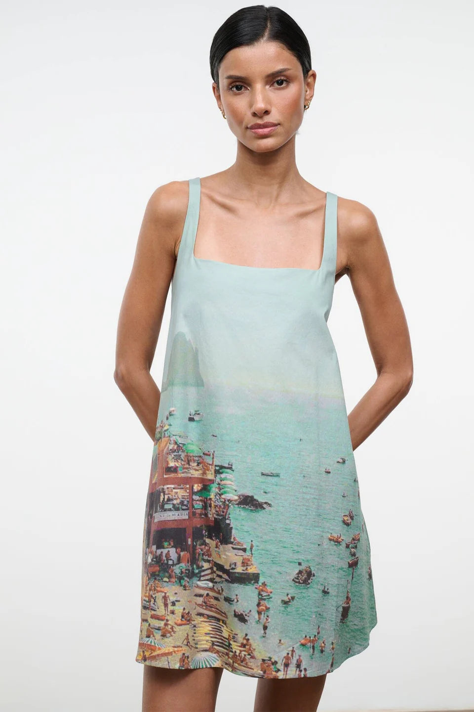 A sleeveless light blue Brio Dress featuring a square neckline and a graphic print of a crowded beach scene.